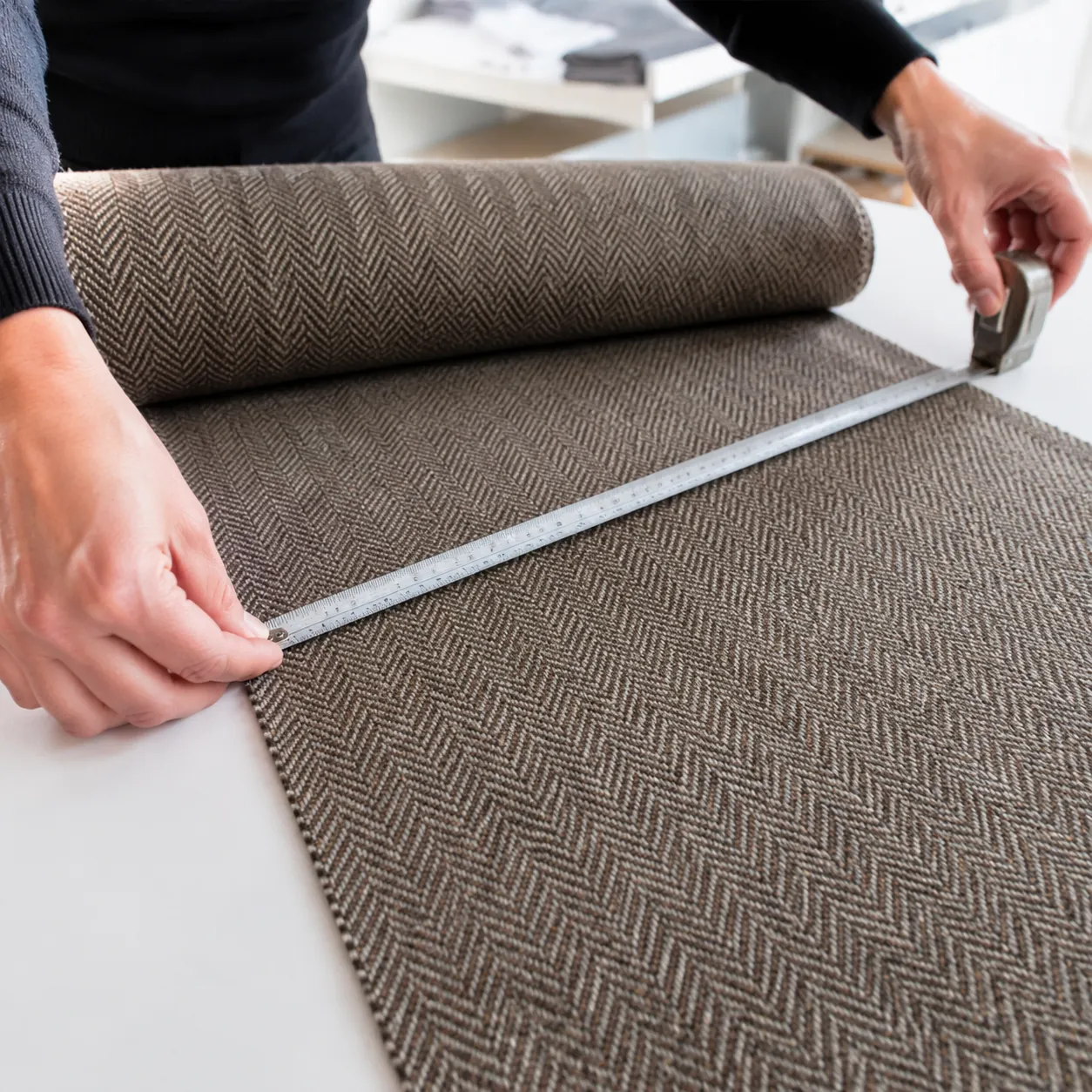 Hyper-realistic product photography, a pair of hands measuring a roll of herringbone tweed fabric with a metal tape measure, bright daylight studio lighting, clean professional workspace background, no text, no brand logo