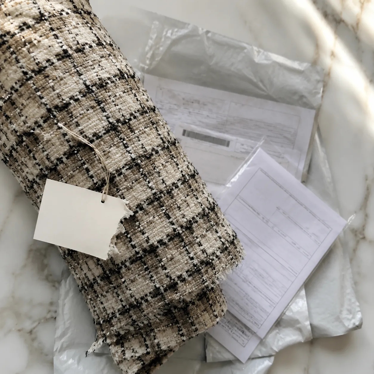 Hyper-realistic product photography, Chanel-style boucl fabric roll with a partially torn retail price tag, overhead view of shipping labels and customs documents, natural window light, no text, no brand logo